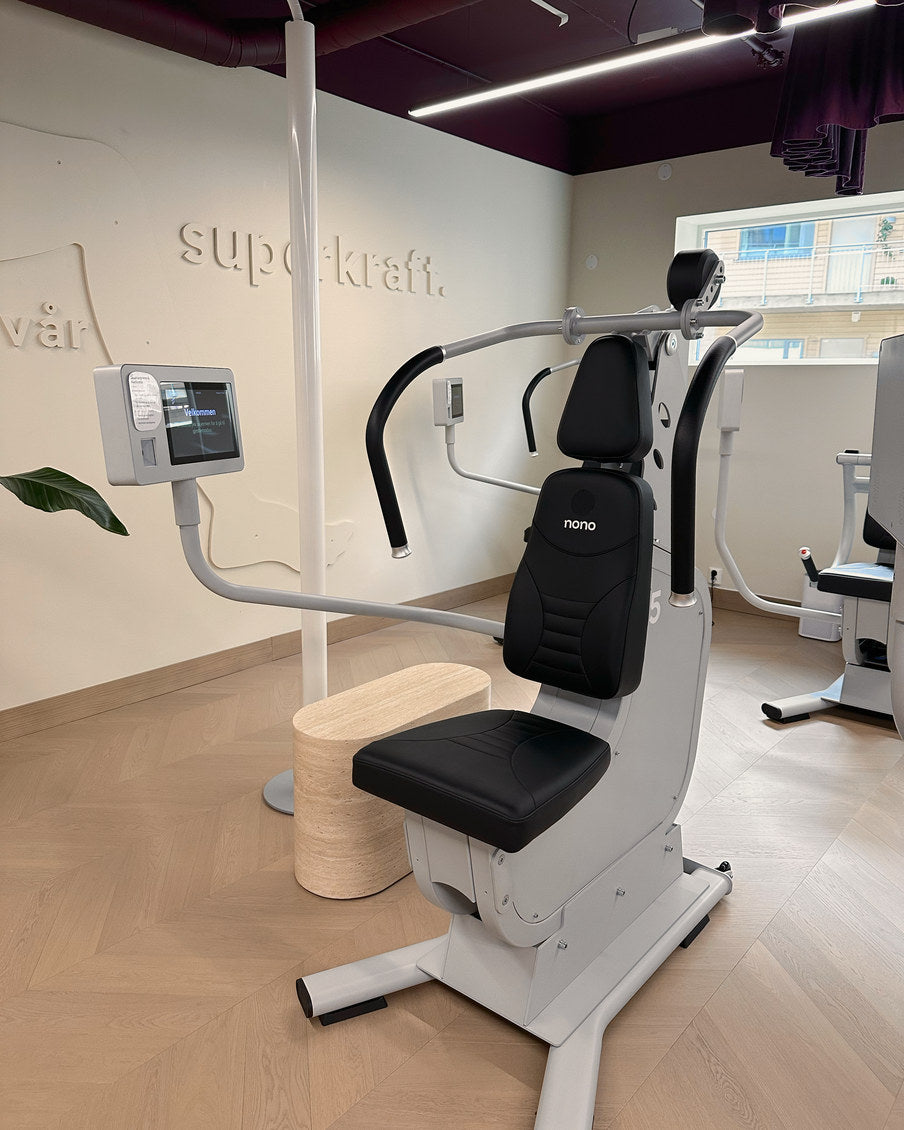 Skulderpress- og nedtrekksmaskinen sett skrått forfra, med skjermen på arm og veggteksten bak.
