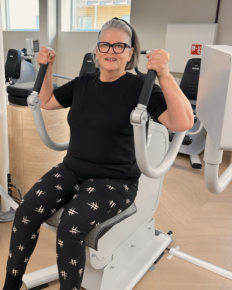 Et medlem i svart t-skjorte bøyer håndtakene opp mot skuldrene i biceps- og tricepsmaskinen og smiler.