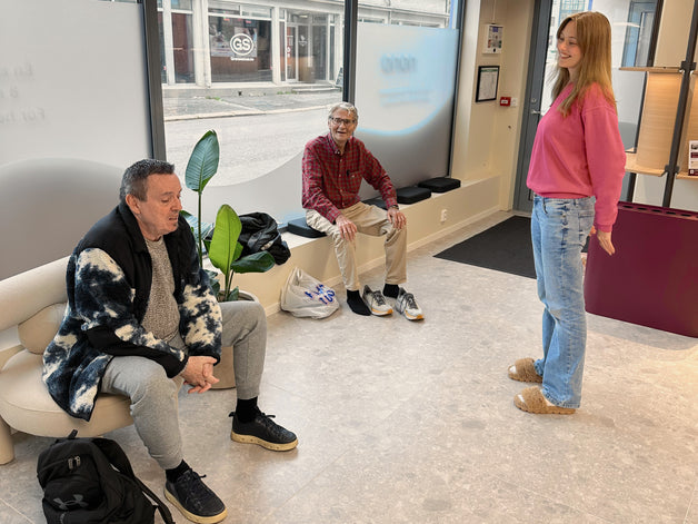 Inngangssonen hos Nono Moss: en vert i rosa genser står og smiler til to medlemmer som sitter ved vinduet mot gata.